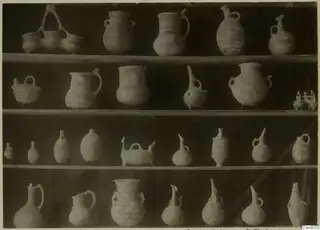 Four shelves of painted pottery jugs, juglets and jars with lattice and diamond patterns, photographed straight on