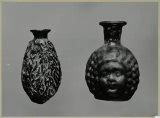 Two blown glass vessels, one with a moulded human face on its body, photographed against a grey background