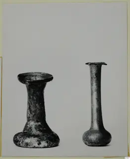 Two small glass vessels side by side on a pale background, one with a flared foot and short neck, one tall and slender-necked.