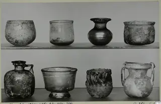 Eight blown glass vessels in two rows on shelves, including small jugs and jars with handles.