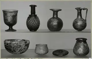 Blown glass vessels on two shelves: a footed cup, bottles, a handled jug, a deep bowl, a small cup and a jar.