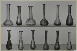 Blown glass vases with long narrow necks, arranged in two rows on shelves against a grey backdrop