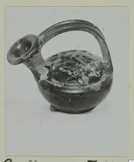 Black glazed vessel with a spout and strap handle, shown on a mounted photograph card