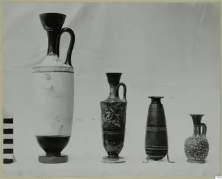 Four vessels in a row on white cloth: a tall white-ground lekythos, a black juglet, a slender banded vessel and a small juglet.