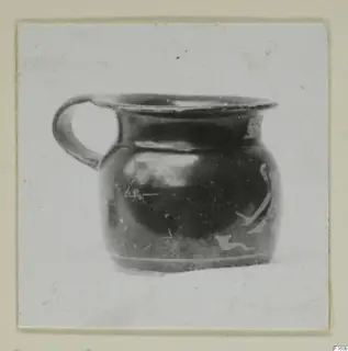 Small jug with a loop handle, dark glossy surface and pale painted motifs on the body, shown on a card mount