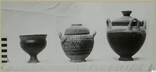Three pottery vessels on a cloth: a footed bowl, a lidded jar with lattice bands, and a two-handled lidded jar.