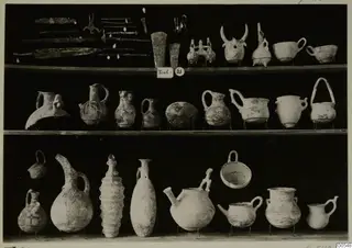 Finds from Lapithos Markides, T.21, on three shelves: metal blades, pins and pottery vessels against a black background.
