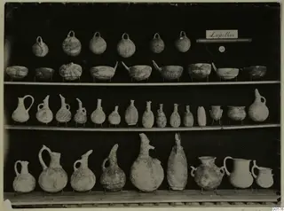 Shelves of pottery vessels from Lapithos Markides, T.49: loop-handled jugs, open bowls and juglets on stands.