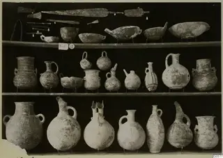 Pottery jugs, bowls and jars with metal blades and pins on three shelves, from T.18 at Lapithos Markides