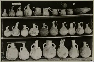 Pottery juglets, jugs and a shallow bowl arranged in three rows on shelves, from Lapithos Markides Tomb 18 A.
