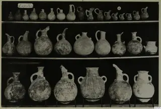 Three shelves of pottery jugs and juglets photographed against a black background