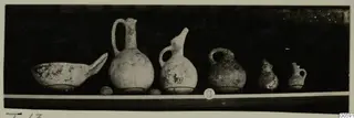 Row of six pottery vessels from Lapithos Markides, T.13, standing on a shelf against a dark background