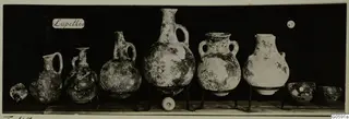 Row of pottery jugs, jars and bowls from Lapithos, Markides tomb 48, set on metal stands against a dark background.