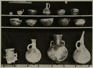 Jugs and bowls from Lapithos Markides, T.45, shown on two shelves and on wire stands against a dark backdrop.