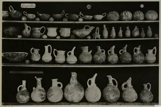 Shelves of pottery jugs, juglets and bowls from Lapithos Markides, T.29, photographed against a black background