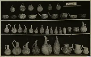 Jars, juglets and bowls from Lapithos Markides T.49, arranged in rows on shelves against a dark background