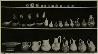 Three shelves of pottery vessels, bowls and jugs from Lapithos Markides, T.49, photographed against a black background.