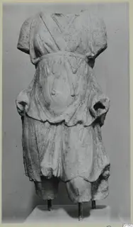 Marble statue of a standing figure, headless and armless, wearing a belted garment, on a metal-supported base.