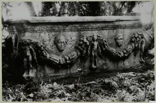 Long side of a stone sarcophagus with carved garland swags and human heads in relief, set among leaves outdoors.