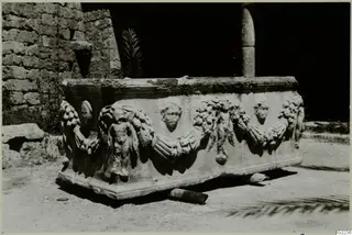 Stone sarcophagus with carved garlands, medallion heads and small figures, standing outdoors by a rubble stone wall.