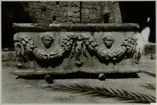 Front of a carved stone sarcophagus with garlands, two carved faces and small figures, standing on dirt before a stone wall
