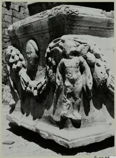 Carved stone block with a head in a round medallion, a garland, and a standing figure in relief.