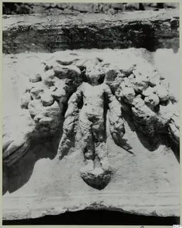 Close view of a carved stone relief showing a small standing figure flanked by scroll-like foliage