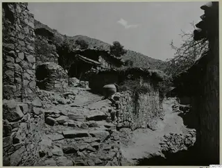 Narrow village lane between stone walls, with rough stone steps on the left and a large clay jar on a wall