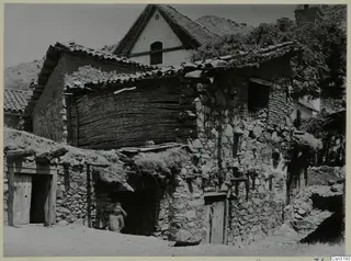 Stone houses with tiled roofs in the village of Lazania, with a stacked reed fence and a person in a doorway