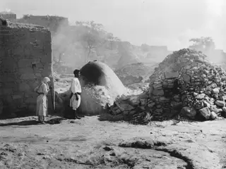 Two men stand beside a domed clay oven and a stone-built dome, with smoke drifting in the background.