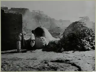 Two people in white robes beside a rounded clay oven and a dome of rough stones, with smoke drifting behind them