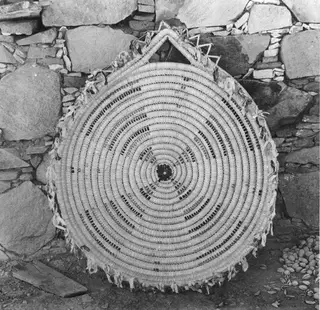 Round coiled handicraft mat with dark spiral stitching, leaning against a dry-stone wall