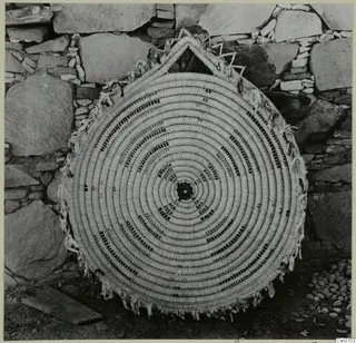 Large circular coiled woven mat with dark patterned bands, leaning against a stone wall