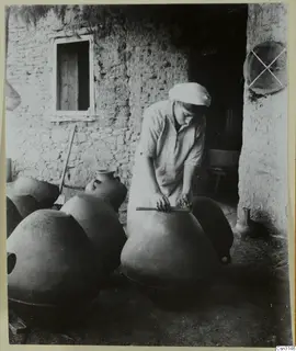 Woman smoothing a large clay jar with a stick outside a stone and mud-walled workshop at Klirou