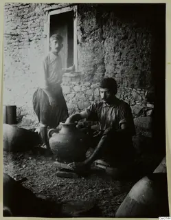 Man seated barefoot working a large clay vessel at Klirou farmstead F, with a second man standing in a doorway behind