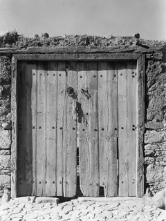 Wooden plank door with studded frame and iron ring knocker in a stone wall, under a thatched roof edge.