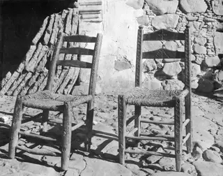 Two wooden ladder-back chairs with woven seats standing against a plastered stone wall.