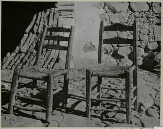 Two wooden chairs with woven seats standing against a stone and plaster wall on a paved ground.