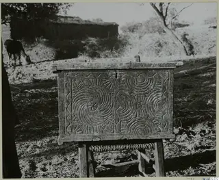 Carved wooden chest with concentric circle and wave patterns, on a wooden stand outdoors beside a mud-walled building.