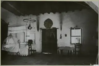 Interior of a village room with a bed, a hanging oil lamp, a wooden door and a barred window