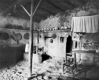 Interior of a village house with plates on a shelf, a row of bottles, hanging woven sieves and a wooden chair