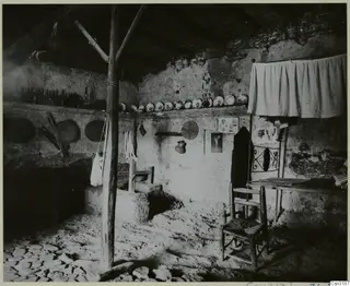Interior of a plastered room with a wooden roof post, a shelf of plates and bottles, and a woven wooden chair.