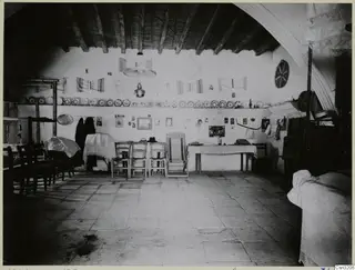 Interior of a whitewashed room with wooden chairs, a table, plates on a wall shelf and striped woven textiles hanging on the wall