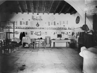 Interior of a vaulted room with chairs, a table, plates on wall shelves and hanging embroidered textiles