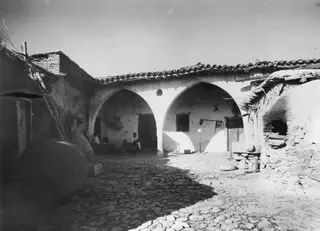 Courtyard at Klirou with arched openings, large storage jars, a leaning ladder and a stone oven at the right edge