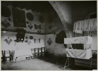 Interior of a room with a tall arch, woven textiles and plates on the walls, a table, chairs and a canopied bed.