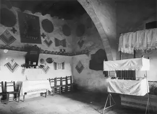 Interior of a vaulted room with textile hangings on the walls, a row of wooden chairs, a covered table and plates on a ledge.