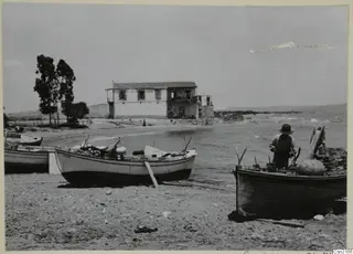 Small boats on a pebble beach at Boghazi, with a two-storey building and a man standing in a boat.