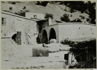 Village houses with arched openings and a wooden ladder, below a tree-covered hillside at Mandres