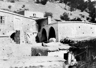 Stone buildings with arched openings and a ladder against a wall, with a wooded hillside behind, at Mandres